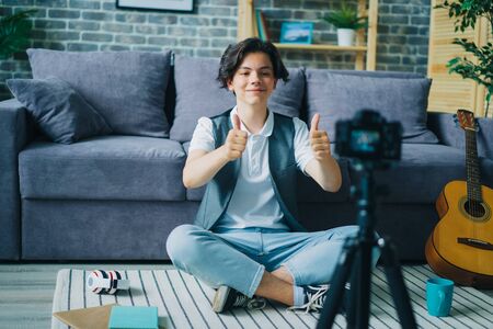Creative boy showing thumbs-up sitting on floor in front of camera recording video for internet vlog. Modern lifestyle, happiness and people concept.の写真素材