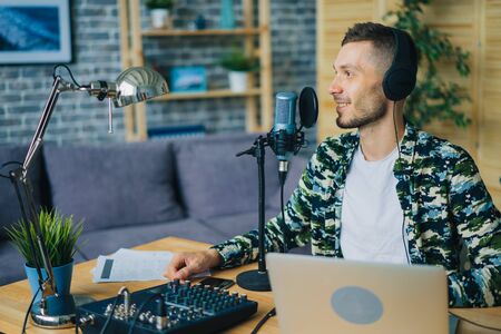 Joyful guy is talking in microphone wearing headphones using laptop in studio sitting at desk alone creating podcast. Modern mass media and equipment concept.の写真素材