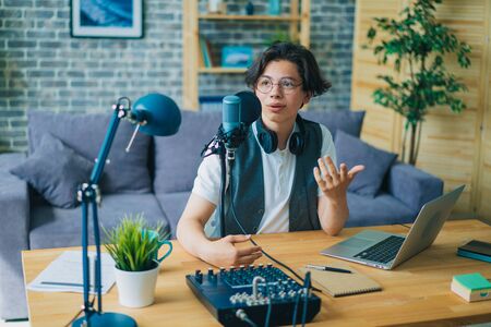 Creative smiling teenage blogger recording podcast audio in microphone sitting at table at home talking in microphone. Youth and blogging concept.の写真素材
