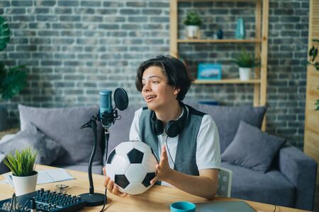 Happy teenager sports fan is talking in microphone in recording studio holding football creating content for internet blog. Youth and modern lifestyle concept.の写真素材