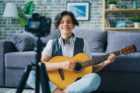 Portrait of teenage blogger recording video at home holding guitar smling using professional camera on tripod. Modern technology and vlogging concept.の写真素材