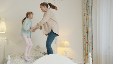 Mother and daughter are jumping in bed having fun holding hands enjoying free time indoors. Family activities, people and modern lifestyle concept.の写真素材