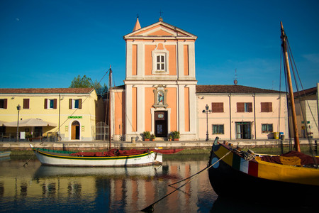 The facade of San Giacomo church, waterfront building in Porto Canale, Cesenatico, Emilia Romagna, Italy. With typical colorful boats.のeditorial素材