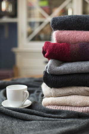 Pile of colorful sweaters with hot drink white cup on the side, background with copy space, cozy hygge wallpaper.の写真素材