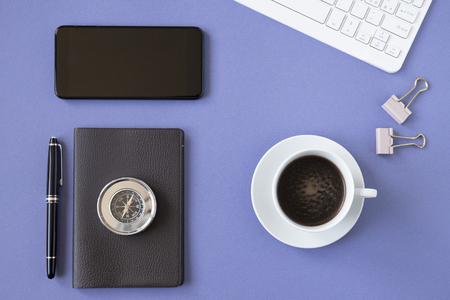 Office desk top view or flat lay with blue background, white keyboard, cup of coffee, notebook with black pen and a metal compass.の写真素材