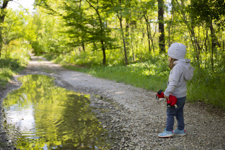 Toddler girl on woodland unpaved road watching a big puddle and holding a red plush toyの写真素材