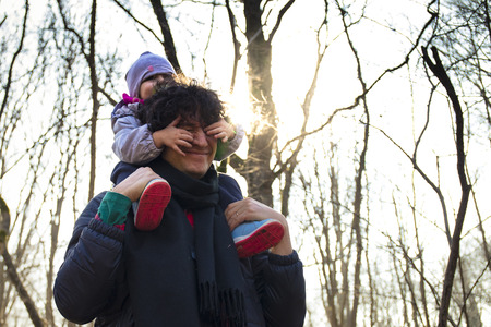 Young dad carrying baby on his shoulders in a forest. Baby covering her dad's eyes for fun.の写真素材