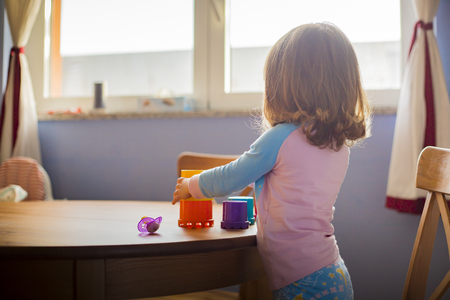 toddler baby girl playing with colorful toys on wooden table  in pink and blue pajamas. Back view, morning light.の写真素材