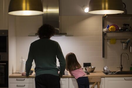 Young father and little child girl, back view, cooking together in modern white kitchen with big brass lampsの写真素材