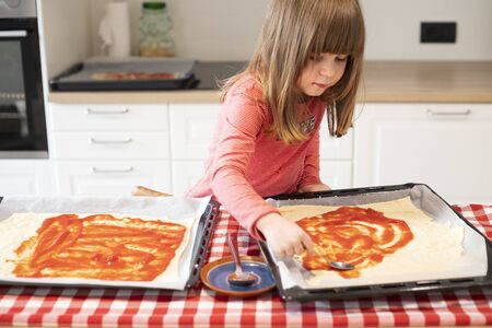 Child girl in a kitchen spreading tomato sauce on raw pizza before baking. Lockdown activity idea for kids. Front view.の写真素材