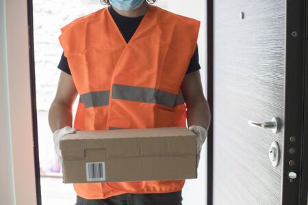 Young delivery man at front door with a parcel box, wearing gloves and face mask during covid-19 pandemic lockdown. Front view.の写真素材