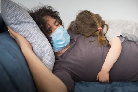 Caucasian father wearing face mask to avoid contagion sleeping with little child girl  during covid-19 pandemic lockdown. Dad and baby daughter sleeping together and embracing.の写真素材