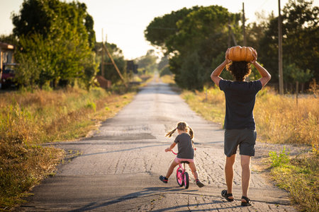Little child girl, riding a bike, with her young father carrying a big halloween pumpkin over his head, on a country road at sunset. Back view.の写真素材