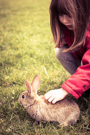 Little Child girl, wearing red jacket, cuddling Easter bunny in the grass. Spring background,...