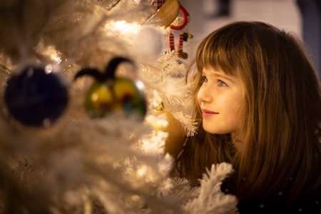 Smiling 5 year old child girl, standing near a white illuminated christmas tree, looking at the decorations.の写真素材