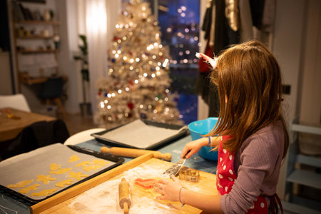Caucasian child girl making Christmas cookies with a christmas tree in the background. Side view.の写真素材