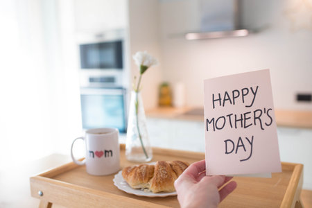 Mother's day still life with mom cup, a sweet pastry and a hand holding a greeting card, in airy indoor background.の写真素材