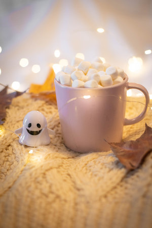 Halloween still life with pink cup filled with chocolate and marshmallows, little ghost and string lights on a white wool sweater. Festive, cozy background with copy space. Vertical shot.の写真素材
