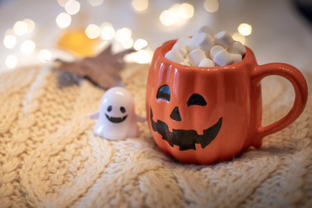 Halloween still life with pumpkin cup filled with chocolate and marshmallows, little ghost and string lights on a white wool sweater. Festive, cozy background with copy space.の写真素材