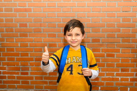 portrait of smiling boy with thumbs-up with student backpack and yellow T-shirt in front of brick...