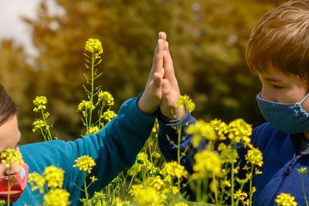 joined hands of two children with face mask in a green field with yellow flowers.