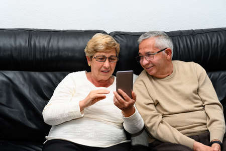 Man and woman looking at the mobile phone. They are sitting on the sofa at homeの写真素材