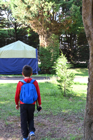 Kid on his back with a blue backpack. The boy is heading for a tent with a walkie talkie in his...