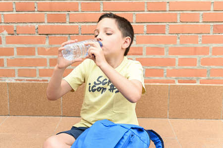 Boy sitting on the ground drinking water from a plastic bottle next to his school bag.の写真素材