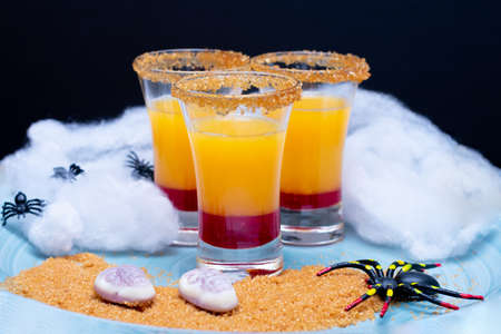 Three glasses with orange juice and grenadine alcohol free surrounded by spiders and halloween candiesの写真素材