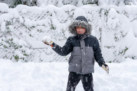 Child standing with a snowball in one hand. Child wears a winter coat with hood over his headの写真素材