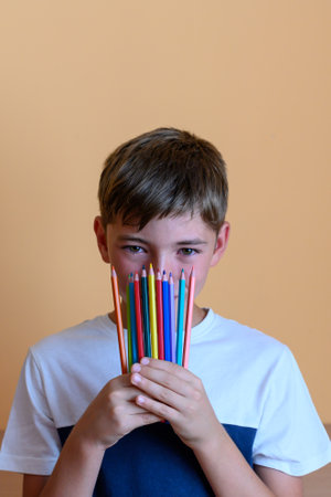 Preteen boy holds crayons covering half of his face on orange background. Back to school conceptの写真素材