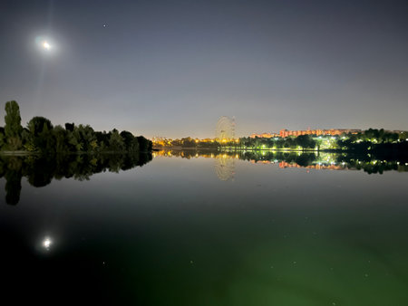 Night view of trees, moon and city reflecting on lake waterの写真素材