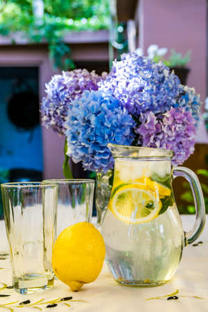 A jug with lemonade, a lemon and empty glasses on a table. Beautiful purple flowers in the background.の写真素材