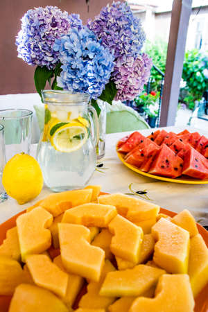 Watermelon triangle slices, yellow orange Mellon slices and lemonade on a table. A vase with flowers in the backgroundの写真素材