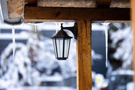 A turned off lantern hanging from a wooden roof. Snow icicles hanging from the rooftop. Snow covered trees in background. High quality photoの写真素材