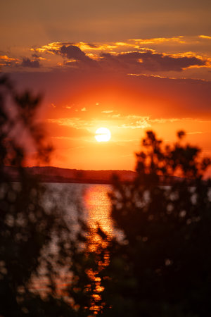 A dramatic orange sunset shot between tree branches and leaves silhouettes. Yellow-red sky with clouds over the Black Sea coast.の写真素材