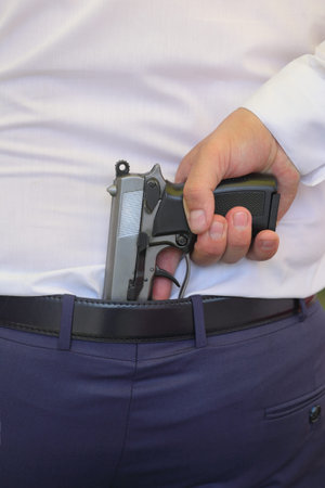 Human hand holding gun behind belt in pants in the back of a man