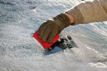 Winter scene, human hand in glove scraping ice from windshield of car, driver using tool