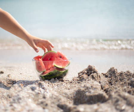 Glass transparent bowl with watermelon slices on the beach. The hand reaches for a refreshing watermelon. Picnic by the ocean sea.の写真素材