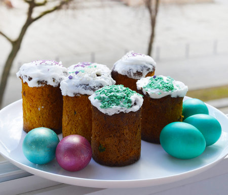 Small Easter cakes and colored eggs on a white plate at the window. Easter in the city.