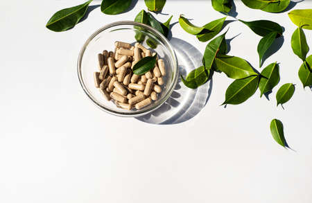 Vitamin capsules in a glass cup. Trace elements and health supplements. Glass shadow and fresh leaves. Copy space for text. white background.の写真素材
