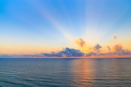 Sunset over a calm ocean casting beams of orange light into the air as the setting sun penetrates the clouds above in a scenic tranquil seascape.の写真素材