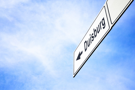 White signboard with an arrow pointing left towards Duisburg, North Rhine-Westphalia, Germany, against a hazy blue sky in a concept of travel, navigation and direction. Path included for the signboardの写真素材