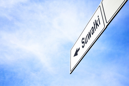 White signboard with an arrow pointing left towards Suwalki, Poland, against a hazy blue sky in a concept of travel, navigation and direction. Path included for the signboardの写真素材
