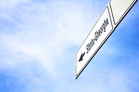 White signboard with an arrow pointing left towards Sfantu-Gheorghe, Romania, against a hazy blue sky in a concept of travel, navigation and direction. Path included for the signboardの写真素材