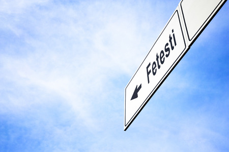 White signboard with an arrow pointing left towards Fetesti, Romania, against a hazy blue sky in a concept of travel, navigation and direction. Path included for the signboardの写真素材