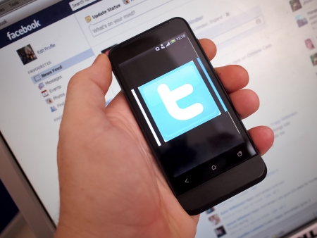 A hand holding a smart phone containing Twitter with a monitor showing Facebook in the backgroundのeditorial素材