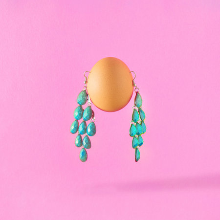 Creative composition of Easter egg with turquoise earrings on a pink background. Minimal holiday or...