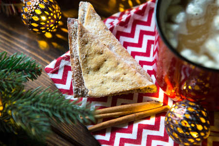 Triangular cookies with powdered sugar and cinnamon on a napkin with a zigzag pattern in the Christmas decor. Red coffee mug with marshmallow, spruce branches, garland, new year, comfort.の写真素材