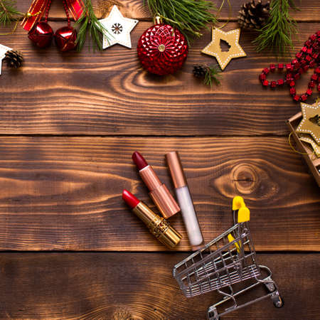 Red lipstick and lip gloss and eyelids on a wooden background in metal supermarket cart in the Christmas decor. Holiday makeup for new year, gift, shopping, women's desires. Space for text, flat layの写真素材